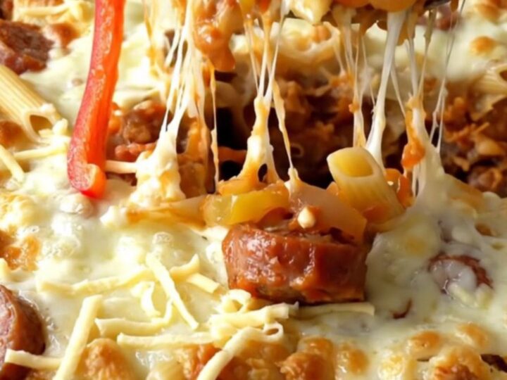 A top-down view of a ceramic baking dish filled with raw penne, sausage coins, bell peppers, and onions.