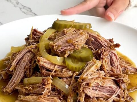 A plate of tender shredded dill pickle pulled pork with visible pickle slices and soft onion pieces.