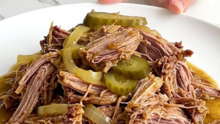 A plate of tender shredded dill pickle pulled pork with visible pickle slices and soft onion pieces.