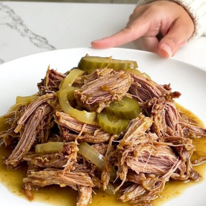 A plate of tender shredded dill pickle pulled pork with visible pickle slices and soft onion pieces.