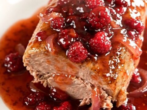 A whole braised pork loin in a slow cooker topped with a glossy raspberry chili sauce and soft red onions.