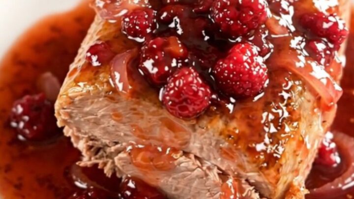 A whole braised pork loin in a slow cooker topped with a glossy raspberry chili sauce and soft red onions.