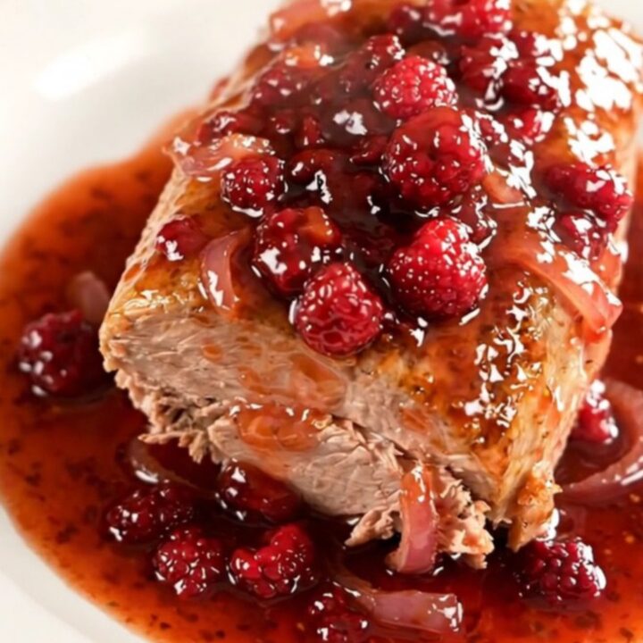 A whole braised pork loin in a slow cooker topped with a glossy raspberry chili sauce and soft red onions.