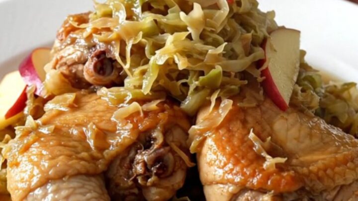 A close-up of bone-in chicken thighs braised with sauerkraut and apple slices on a white plate.