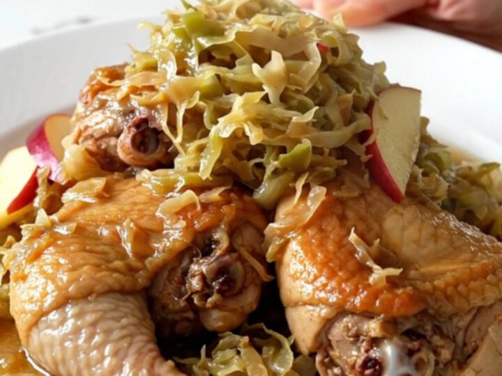 A close-up of bone-in chicken thighs braised with sauerkraut and apple slices on a white plate.