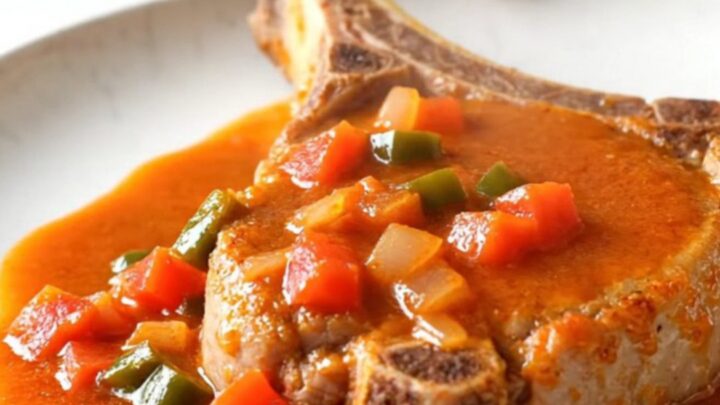 A close-up of a tender bone-in pork chop topped with a zesty ranchero tomato and pepper sauce on a white plate.