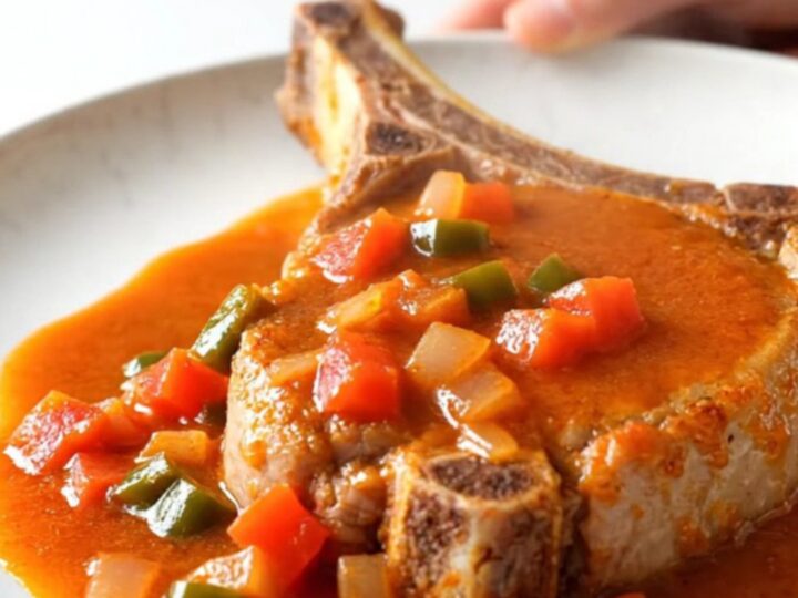 A close-up of a tender bone-in pork chop topped with a zesty ranchero tomato and pepper sauce on a white plate.