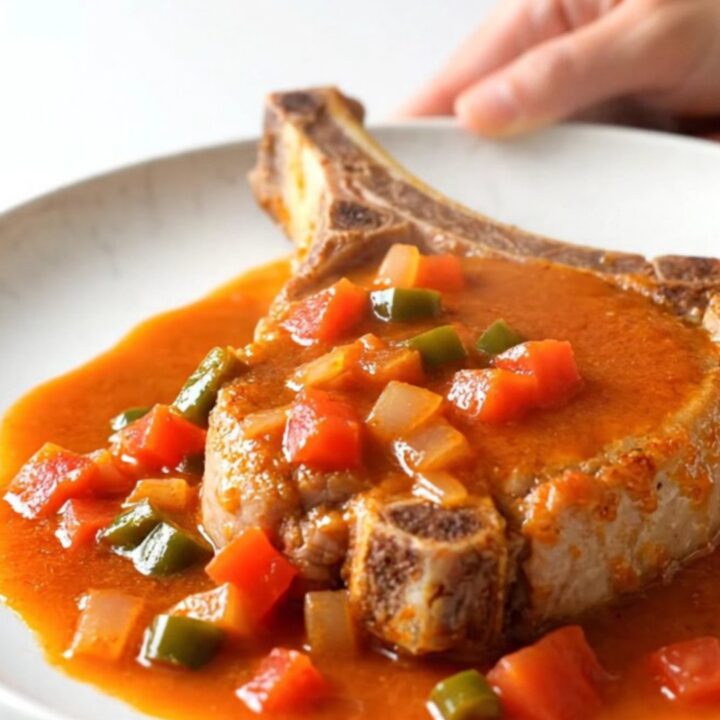 A close-up of a tender bone-in pork chop topped with a zesty ranchero tomato and pepper sauce on a white plate.