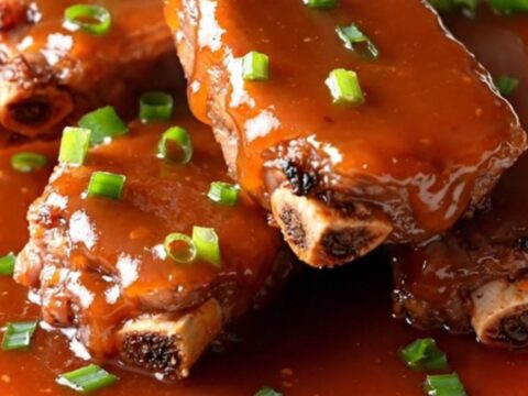 A slow cooker filled with raw pork spareribs being seasoned with green onions and sweet chili sauce.