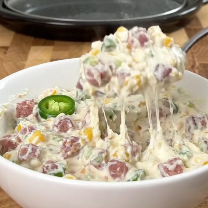 A stainless steel slow cooker filled with a creamy dip featuring sausage, corn, and jalapenos.