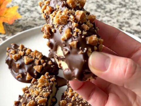A close-up of chocolate-dipped green apple wedges topped with toffee bits on a parchment-lined tray.