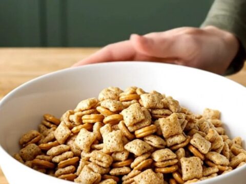A stainless steel slow cooker filled with a golden, toasted mixture of cereal, pretzels, and peanuts.