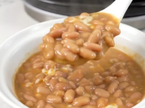 A top-down view of a black slow cooker filled with pinto beans, tomatoes, and cheese cubes.