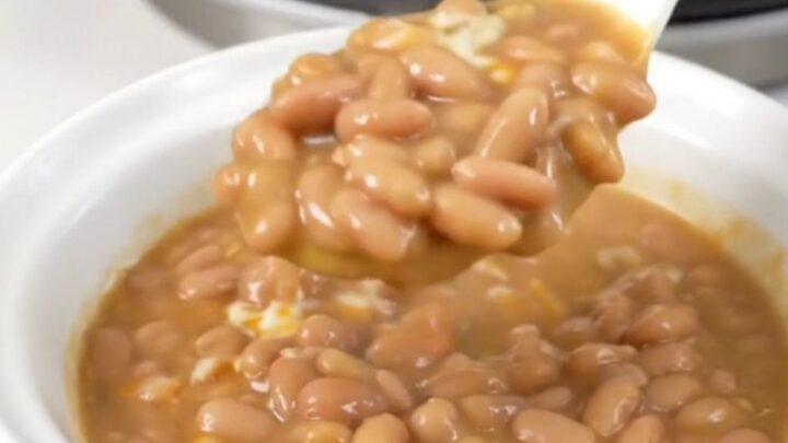 A top-down view of a black slow cooker filled with pinto beans, tomatoes, and cheese cubes.