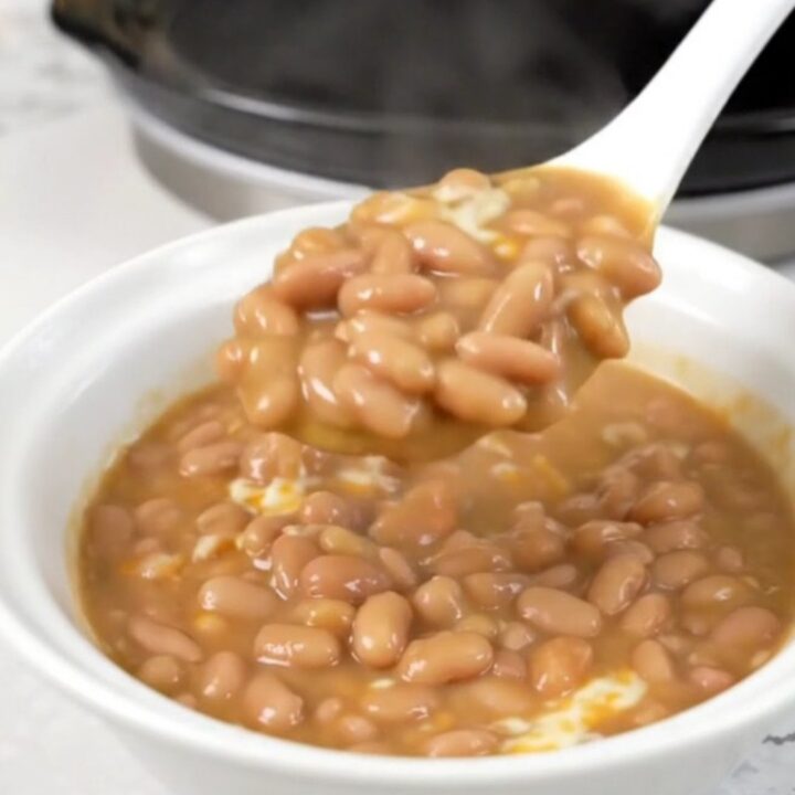 A top-down view of a black slow cooker filled with pinto beans, tomatoes, and cheese cubes.