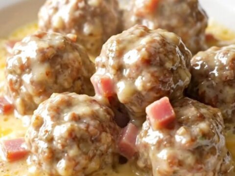 A close-up of tender chicken meatballs in a creamy Swiss cheese and ham sauce inside a black slow cooker.