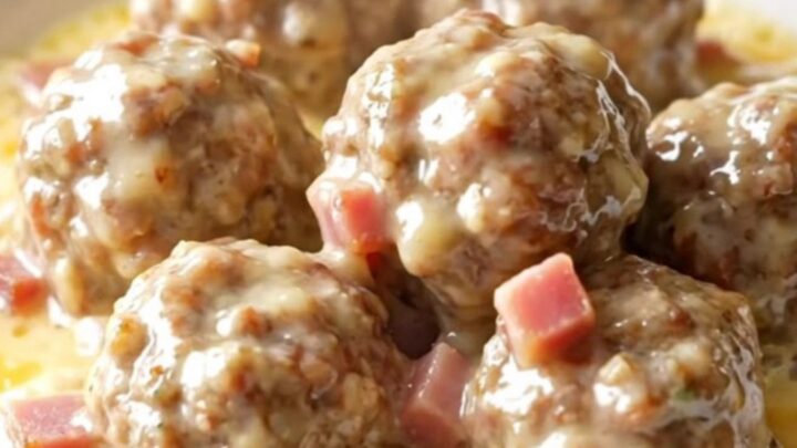 A close-up of tender chicken meatballs in a creamy Swiss cheese and ham sauce inside a black slow cooker.