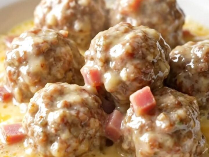 A close-up of tender chicken meatballs in a creamy Swiss cheese and ham sauce inside a black slow cooker.