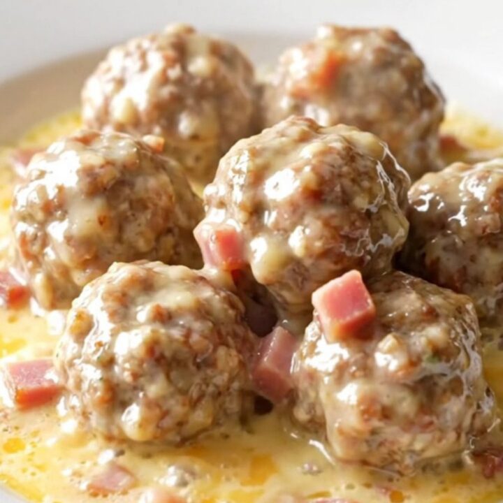A close-up of tender chicken meatballs in a creamy Swiss cheese and ham sauce inside a black slow cooker.