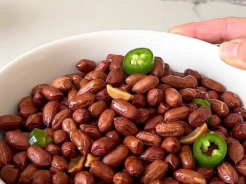 A top-down view of raw peanuts being added to a black slow cooker filled with Cajun spices and jalapeno rings.