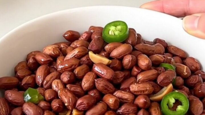 A top-down view of raw peanuts being added to a black slow cooker filled with Cajun spices and jalapeno rings.