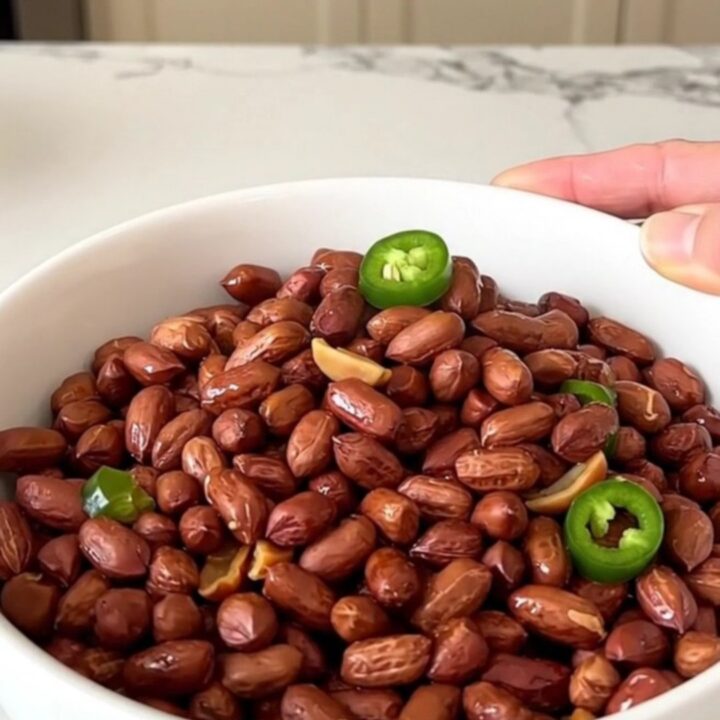 A top-down view of raw peanuts being added to a black slow cooker filled with Cajun spices and jalapeno rings.