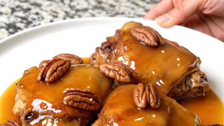A close-up of tender, braised chicken thighs in a rich maple-butter sauce topped with toasted pecans in a slow cooker.
