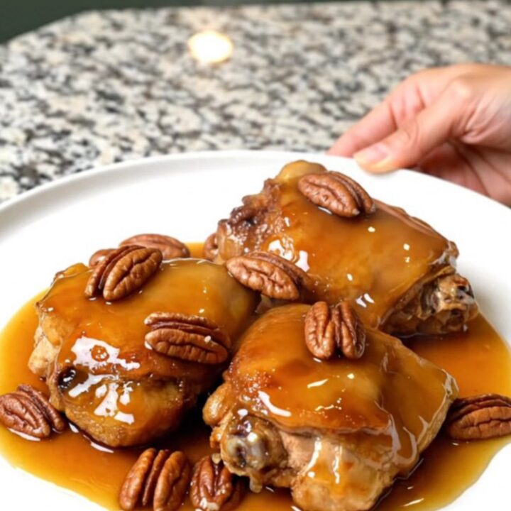 A close-up of tender, braised chicken thighs in a rich maple-butter sauce topped with toasted pecans in a slow cooker.