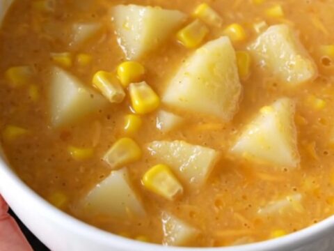 A steaming bowl of creamy potato and corn chowder topped with melted cheddar cheese on a beige background.
