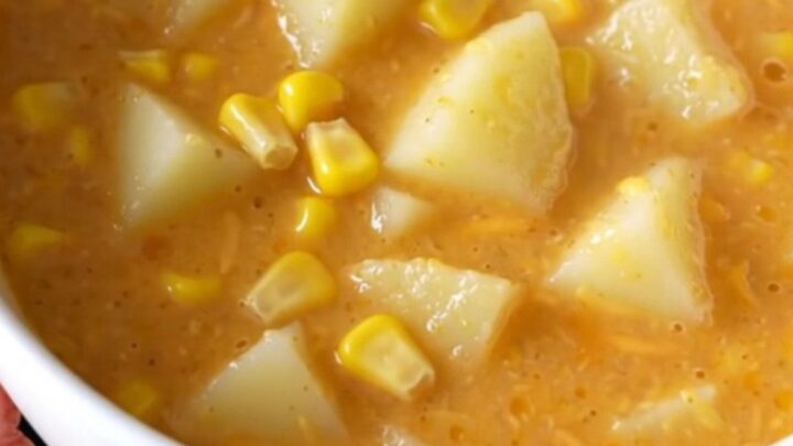 A steaming bowl of creamy potato and corn chowder topped with melted cheddar cheese on a beige background.