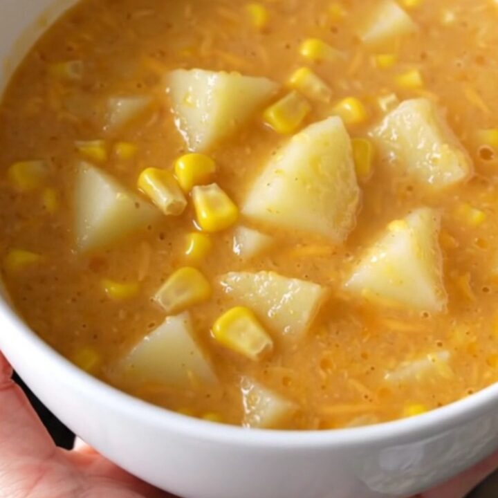 A steaming bowl of creamy potato and corn chowder topped with melted cheddar cheese on a beige background.