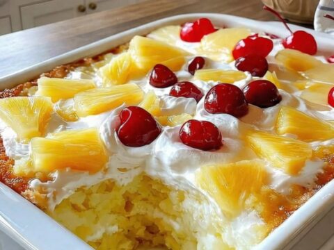 A close-up of a slice of yellow cake topped with whipped cream, pineapple, and a cherry.