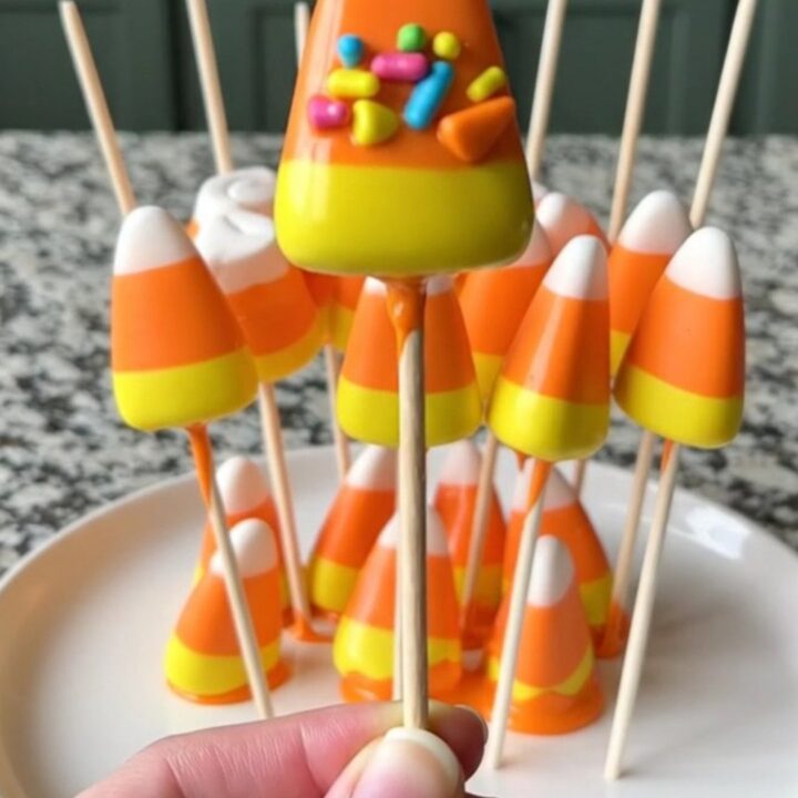 A dozen candy corn marshmallow pops on a baking sheet, featuring bright orange coating and festive candy corn sprinkles.