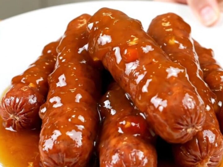 A stainless steel slow cooker filled with mini smoked sausages covered in a glossy amber pepper jelly glaze and diced red