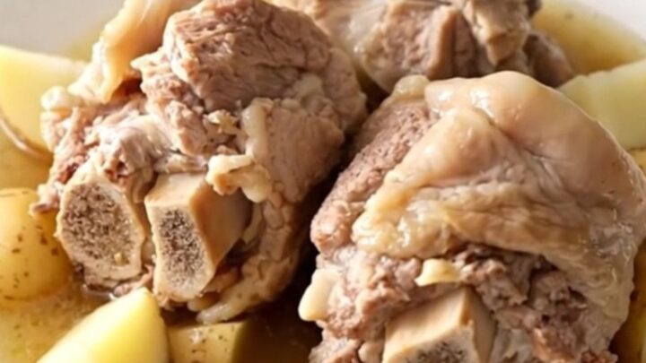 A close-up of tender, braised pork neck bones served with soft potatoes in a rich, savory broth on a white plate.