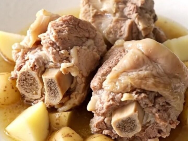 A close-up of tender, braised pork neck bones served with soft potatoes in a rich, savory broth on a white plate.