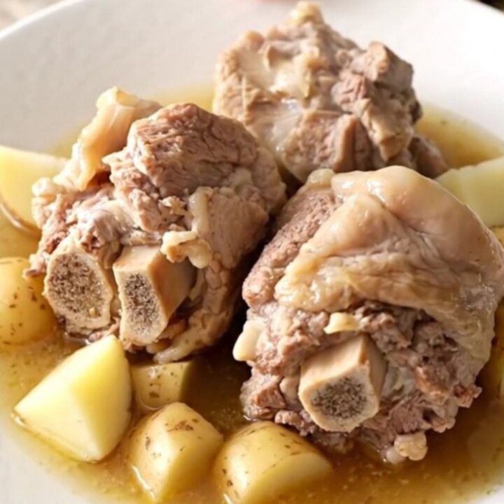 A close-up of tender, braised pork neck bones served with soft potatoes in a rich, savory broth on a white plate.