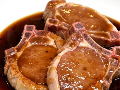Five raw bone-in pork chops fanned out in a black slow cooker insert with garlic and bay leaves.