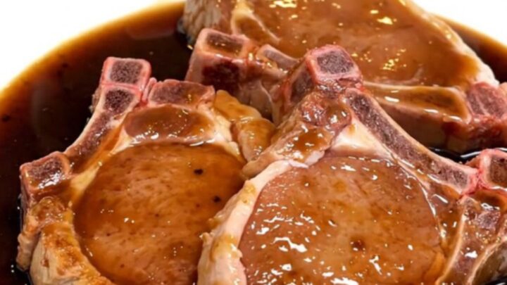 Five raw bone-in pork chops fanned out in a black slow cooker insert with garlic and bay leaves.
