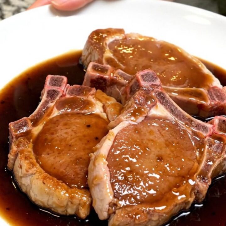 Five raw bone-in pork chops fanned out in a black slow cooker insert with garlic and bay leaves.