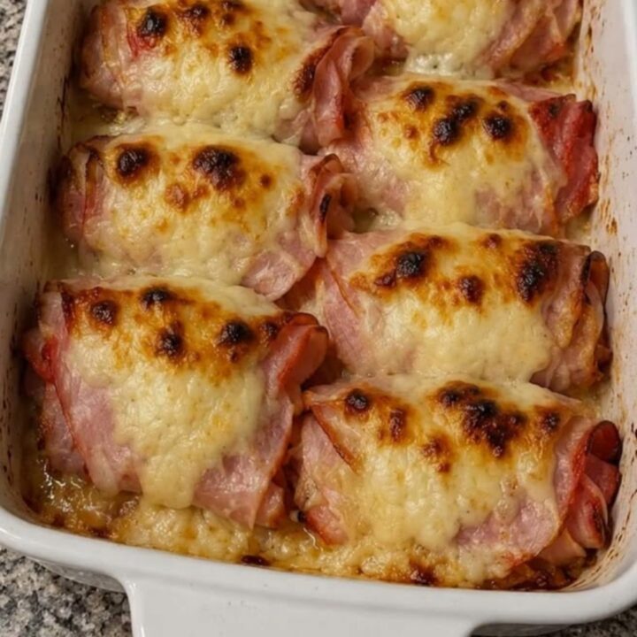 A white casserole dish filled with eight golden-brown, melted cheese-topped ham rolls on a granite countertop.