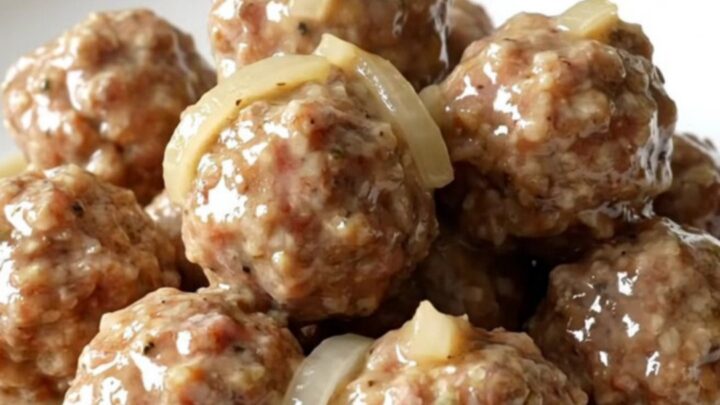 A slow cooker filled with turkey meatballs, sliced onions, and creamy country gravy on a white speckled countertop.
