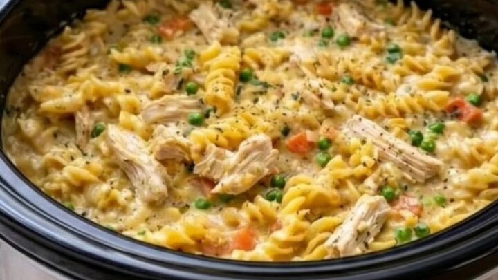 A stainless steel slow cooker filled with creamy rotini pasta, tender shredded chicken, and bright green peas.