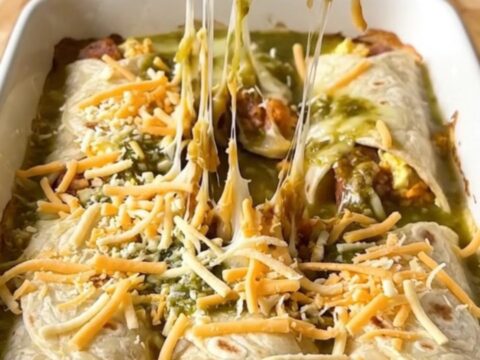 A top-down view of a rectangular baking dish filled with sausage and egg burritos covered in salsa verde and melted pepper