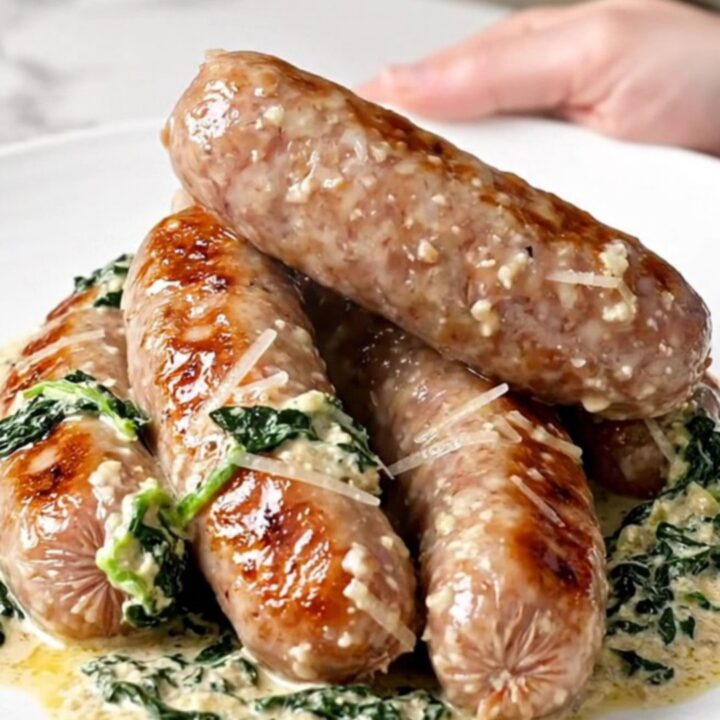 A close-up view of creamy Italian sausage links in a slow cooker topped with wilted spinach and parmesan cheese.