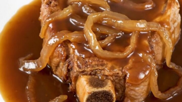 A close-up of tender, slow-cooked bone-in pork steaks covered in a rich brown sugar bourbon sauce with soft onions on a white