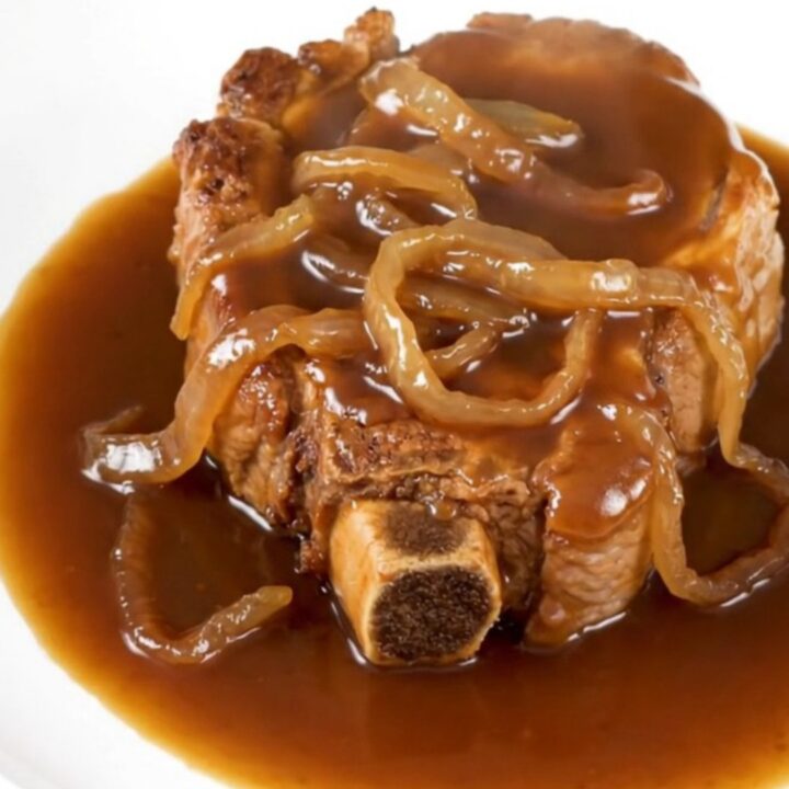 A close-up of tender, slow-cooked bone-in pork steaks covered in a rich brown sugar bourbon sauce with soft onions on a white