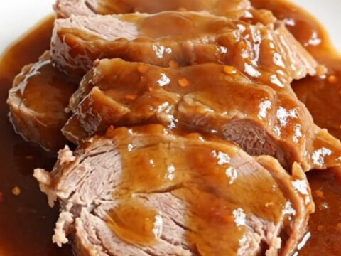 A whole, raw pork tenderloin resting in a slow cooker insert next to a hand adding a second piece.