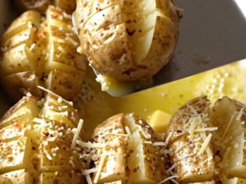 A top-down view of raw, accordion-cut russet potatoes in a white ceramic dish being drizzled with garlic butter.
