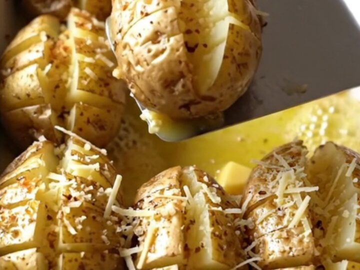 A top-down view of raw, accordion-cut russet potatoes in a white ceramic dish being drizzled with garlic butter.
